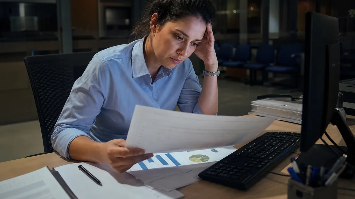 Bancária revisa relatórios de metas em agência com computador aceso ao entardecer