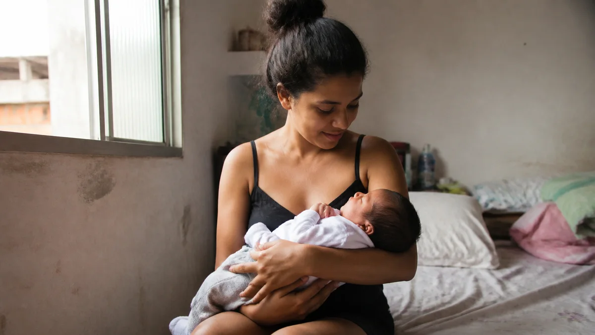 Mãe sentada em quarto simples no Brasil segura recém-nascido no colo sob luz suave da janela