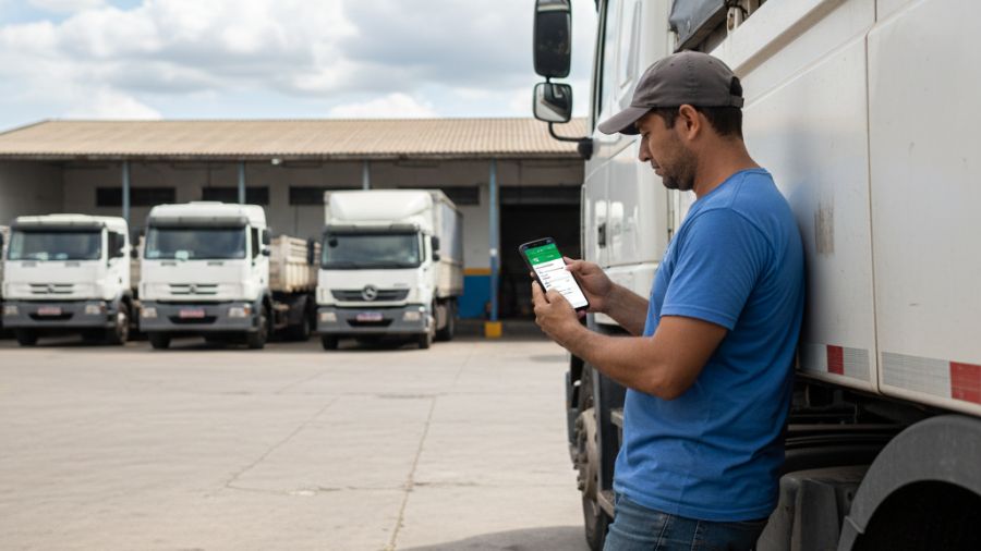 Motorista de caminhão encostado no veículo consultando andamento de processo trabalhista pelo celular em pátio de transportadora